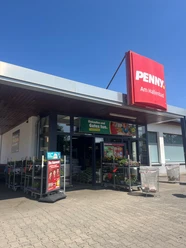 Penny Hallenbad verkleinert.jpg Eingang eines Penny-Supermarkts mit rotem Schild „PENNY. Am Hallenbad“. Vor dem Eingang stehen Einkaufswagen, Blumen auf Rollwagen und Werbeplakate. Der Himmel ist klar und blau.