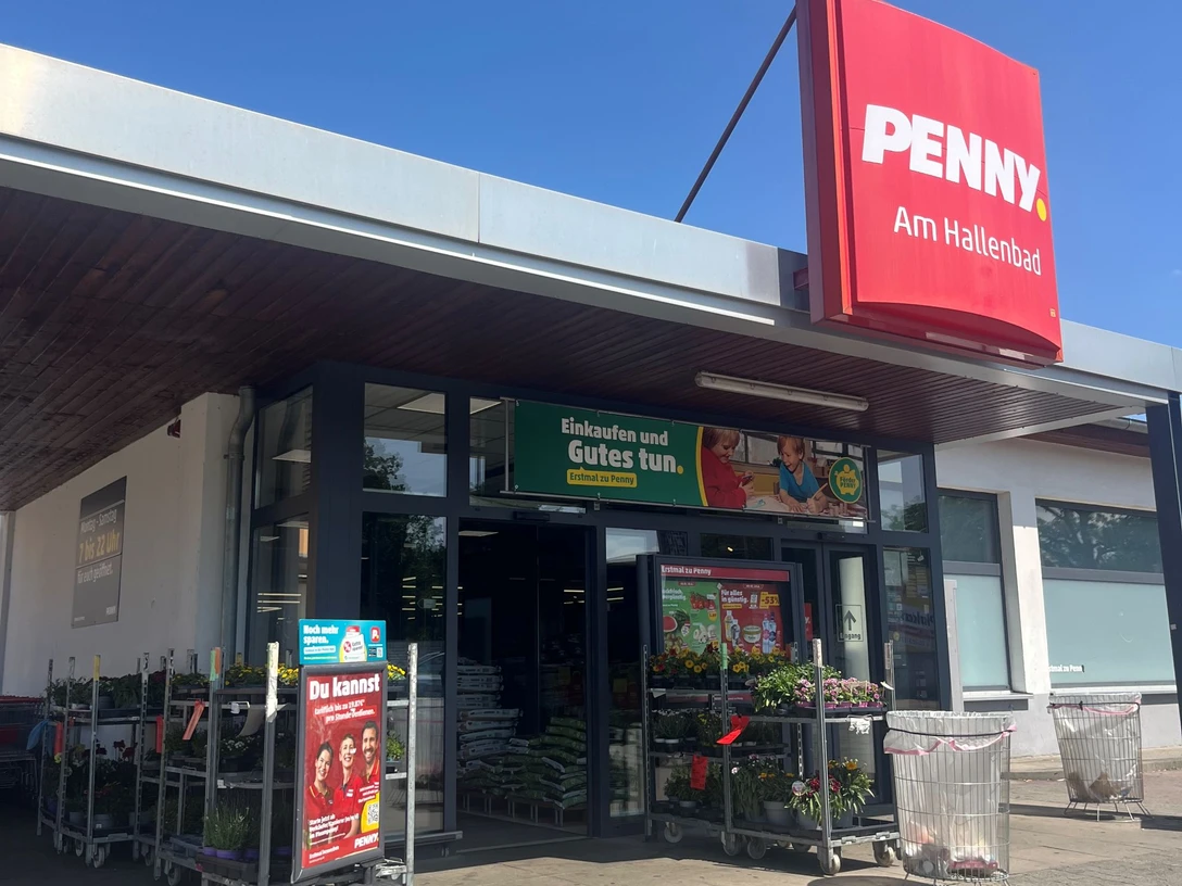 Penny Hallenbad verkleinert.jpg Eingang eines Penny-Supermarkts mit rotem Schild „PENNY. Am Hallenbad“. Vor dem Eingang stehen Einkaufswagen, Blumen auf Rollwagen und Werbeplakate. Der Himmel ist klar und blau.
