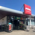Penny Hallenbad verkleinert.jpg Eingang eines Penny-Supermarkts mit rotem Schild „PENNY. Am Hallenbad“. Vor dem Eingang stehen Einkaufswagen, Blumen auf Rollwagen und Werbeplakate. Der Himmel ist klar und blau.