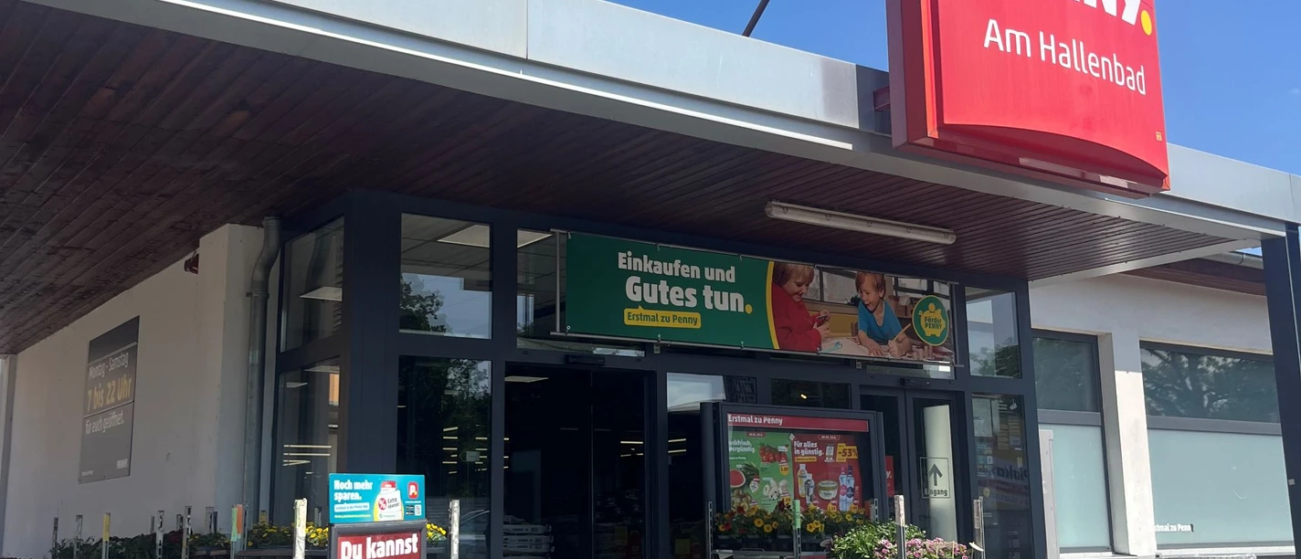 Penny Hallenbad verkleinert.jpg Eingang eines Penny-Supermarkts mit rotem Schild „PENNY. Am Hallenbad“. Vor dem Eingang stehen Einkaufswagen, Blumen auf Rollwagen und Werbeplakate. Der Himmel ist klar und blau.