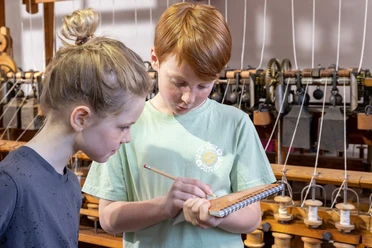 Familienführung Kritzelblock im LVR-Industriemuseum Textilfabrik Cromford, Ratingen Zwei Kinder stehen im LVR-Industriemuseum Cromford mit einem Notizblock vor historischen Webstühlen.