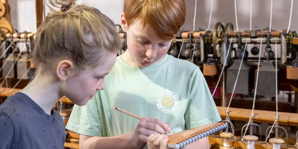 Familienführung Kritzelblock im LVR-Industriemuseum Textilfabrik Cromford, Ratingen Zwei Kinder stehen im LVR-Industriemuseum Cromford mit einem Notizblock vor historischen Webstühlen.