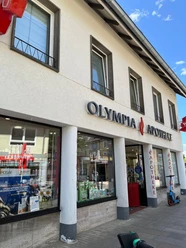 Olympia Apothke vergrößert.jpg Die Olympia Apotheke zeigt eine moderne Fassade mit großen Schaufenstern, beschriftet mit einem auffälligen Logo.