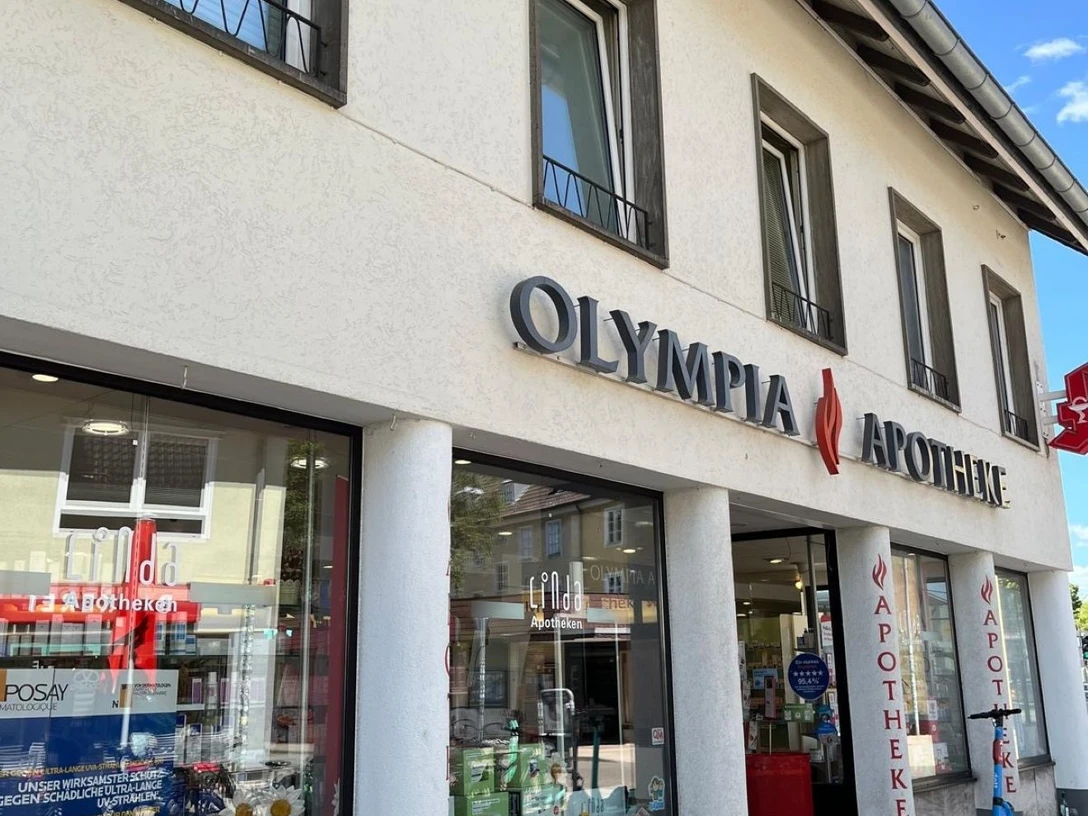 Olympia Apothke vergrößert.jpg Die Olympia Apotheke zeigt eine moderne Fassade mit großen Schaufenstern, beschriftet mit einem auffälligen Logo.