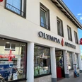 Olympia Apothke vergrößert.jpg Die Olympia Apotheke zeigt eine moderne Fassade mit großen Schaufenstern, beschriftet mit einem auffälligen Logo.