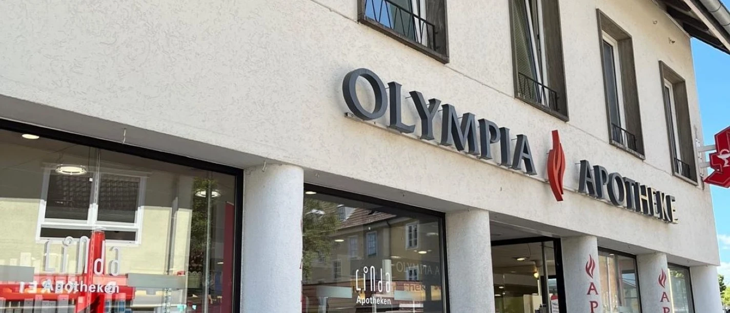 Olympia Apothke vergrößert.jpg Die Olympia Apotheke zeigt eine moderne Fassade mit großen Schaufenstern, beschriftet mit einem auffälligen Logo.