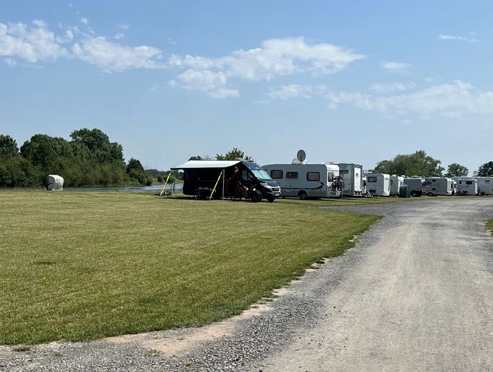 WoMo Weser-Ufer-Urlaub Wohnmobilstellplatz an der Weser mit mehreren Wohnmobilen auf grünem Gras und Schotterweg bei blauem Himmel.