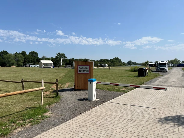 Parkplatz am Weserufer mit Schrankenanlage, Kassenautomat und Wohnmobilen im Hintergrund bei Sonnenschein.
