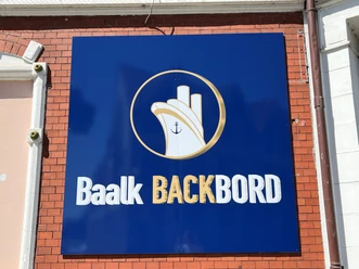 Baalk Backbord_1 Schild an einer Ziegelwand mit maritimem Logo und Schriftzug "Baalk Backbord" in Weiß auf Blau.