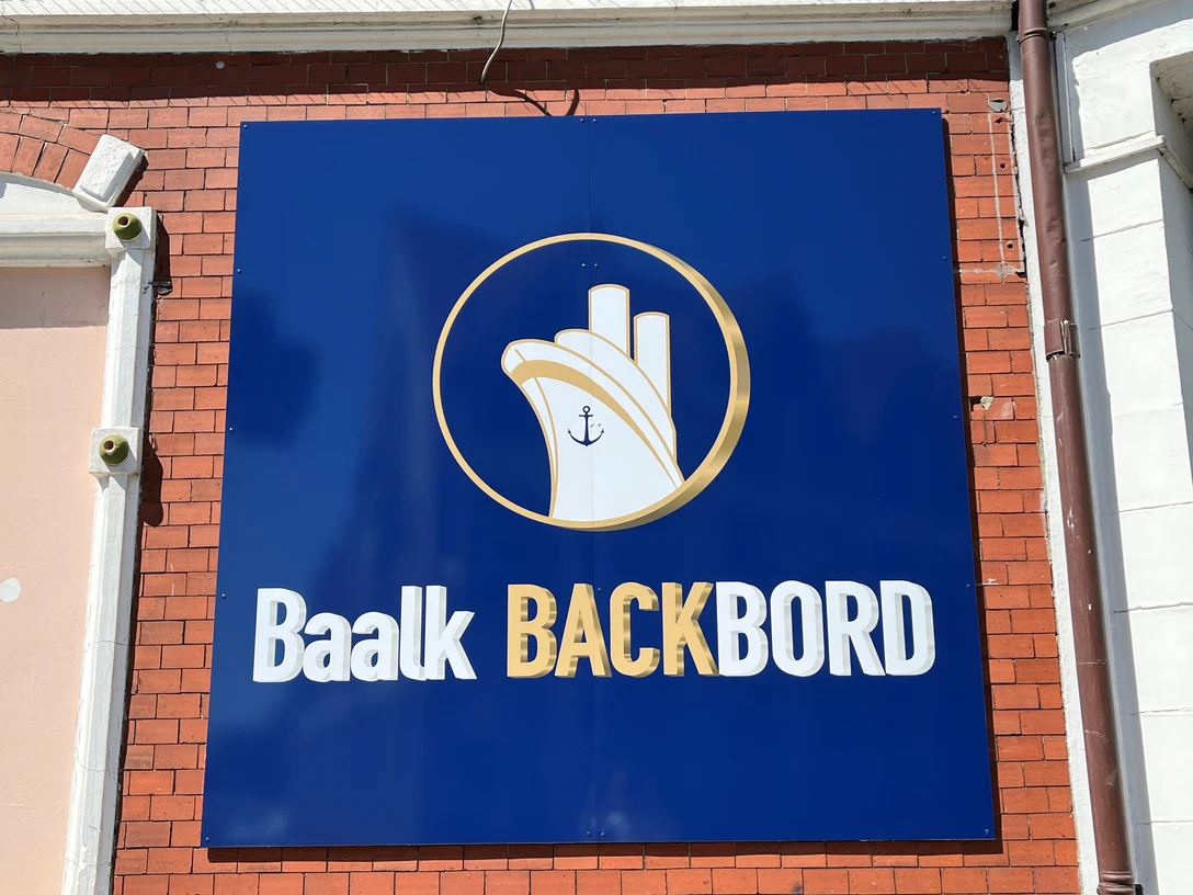 Baalk Backbord_1 Schild an einer Ziegelwand mit maritimem Logo und Schriftzug "Baalk Backbord" in Weiß auf Blau.