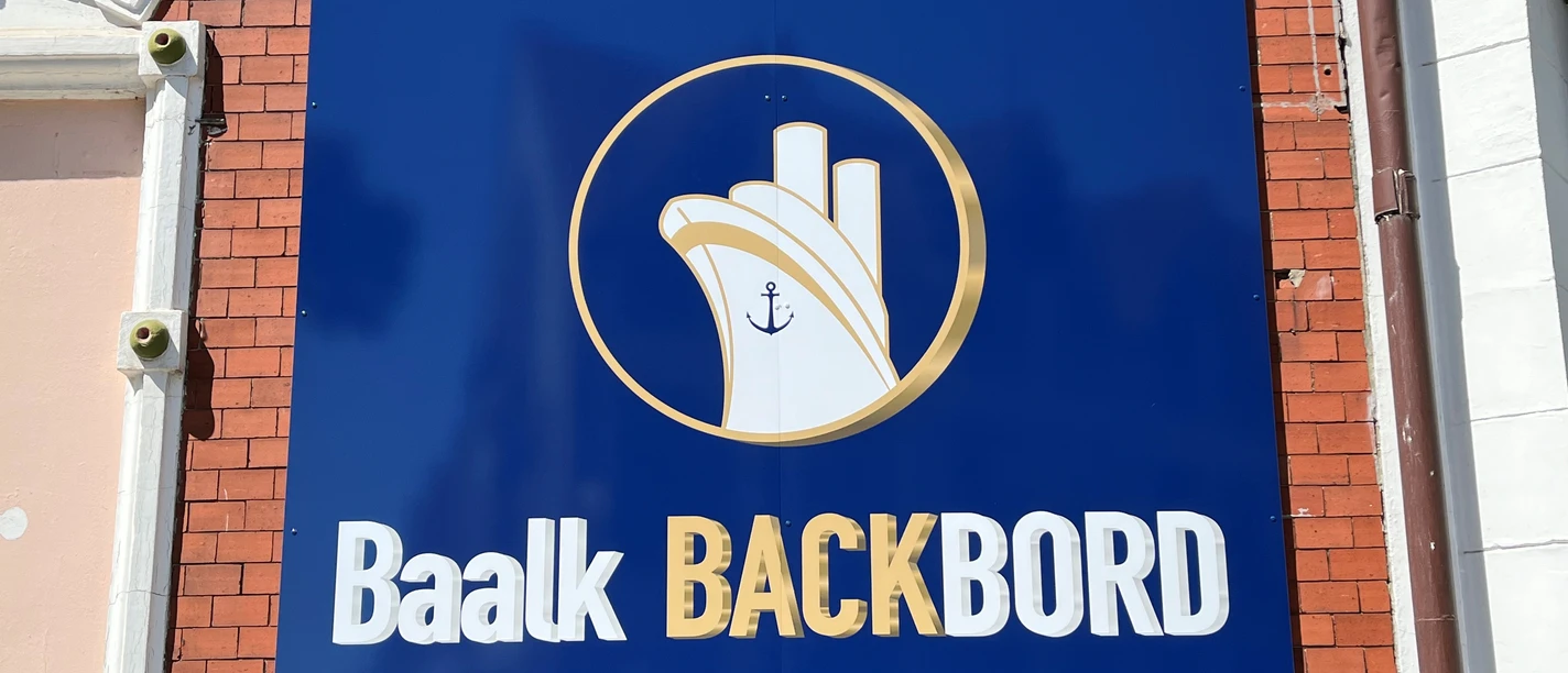 Baalk Backbord_1 Schild an einer Ziegelwand mit maritimem Logo und Schriftzug "Baalk Backbord" in Weiß auf Blau.