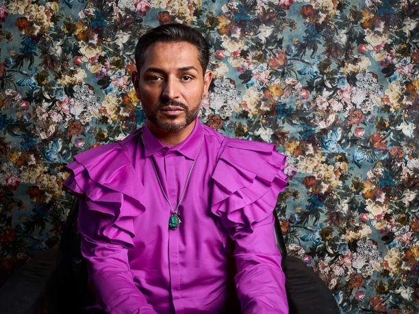 Jovanoic, Gianni A person in a bright pink shirt with ruffles sits in front of a floral patterned wallpaper.