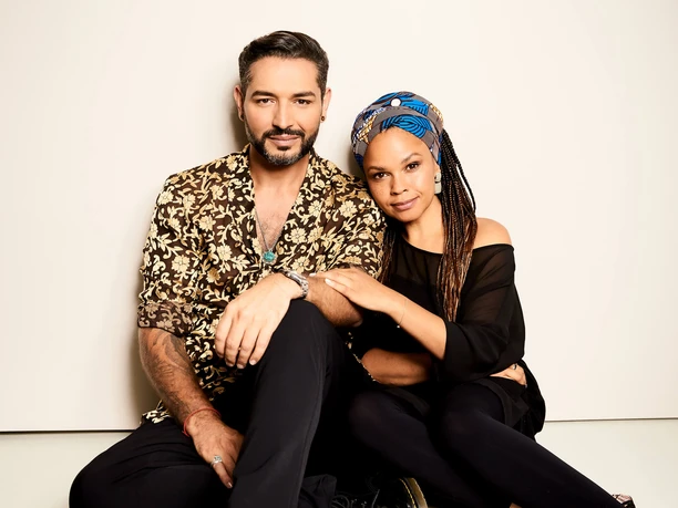Jovanovic, Gianni Oyindamola Alashe A smiling man and a woman sit leaning against each other in front of a neutral background.