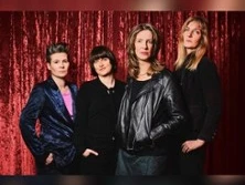 Vier Frauen stehen nebeneinander vor einem roten Bühnenvorhang, tragen stilvolle, dunkle Kleidung.