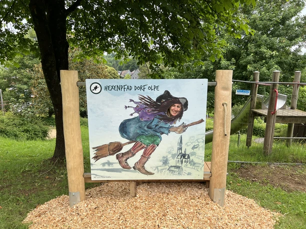 Hexenpfad Dorf Olpe Holztafel im Grünen zeigt fröhliche Hexe auf Besen in buntem Gewand, Teil des Hexenpfads Olpe.