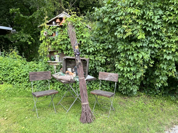 Hexenpfad Dorf Olpe Garten mit zwei Holzstühlen, einem Tisch, dekorativen Pflanzentöpfen und grüner Blatthecke im Hintergrund.