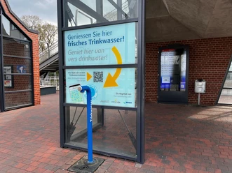 Trinkwasserspender am Bahnhof.jpg Ein moderner Trinkwasserspender steht auf einem Bahnsteig; umgeben von Glaswänden und Backsteinstrukturen strahlt er Frische aus.