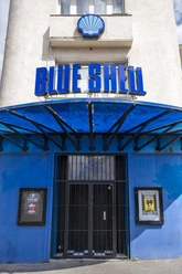 Blue Shell The image shows the entrance area of the venue "Blue Shell" in Cologne. Above the entryway is a prominent blue sign featuring the inscription "Blue Shell." The building has a light-colored facade, while the entry area is accented in blue. A metal canopy provides weather protection and emphasizes the contemporary design of the club.