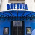 Blue Shell Das Bild zeigt den Eingangsbereich des Veranstaltungsortes „Blue Shell“ in Köln. Über dem Eingang prangt ein auffälliges, blaues Schild mit dem Schriftzug „Blue Shell“. Das Gebäude hat eine helle Fassade, wobei der Eingangsbereich in Blau gehalten ist. Ein Vordach aus Metall bietet Schutz vor Witterungseinflüssen und unterstreicht das zeitgemäße Design des Clubs.The image shows the entrance area of the venue "Blue Shell" in Cologne. Above the entryway is a prominent blue sign featuring the inscription "Blue Shell." The building has a light-colored facade, while the entry area is accented in blue. A metal canopy provides weather protection and emphasizes the contemporary design of the club.