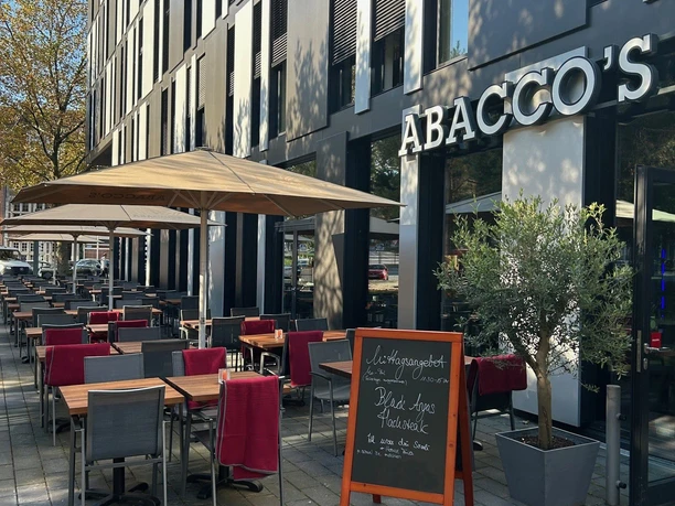 Abaccos Steakhouse Außen_1.jpg Moderne Vorderfront und Eingang zum Abacco's Steakhouse mit großen Fenstern und einer Terrasse mit Stühlen und roten Decken. Auf einer Kreidetafel steht das Mittagsmenü.