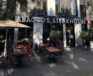 Vorderfront und Eingang des Abacco's Steakhouse mit großen Fenstern und einer Terrasse mit Stühlen und roten Decken.