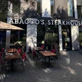 Abaccos Steakhouse Außen_2.jpg Vorderfront und Eingang des Abacco's Steakhouse mit großen Fenstern und einer Terrasse mit Stühlen und roten Decken.