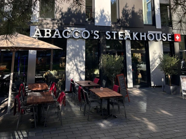 Abaccos Steakhouse Außen_2.jpg Vorderfront und Eingang des Abacco's Steakhouse mit großen Fenstern und einer Terrasse mit Stühlen und roten Decken.
