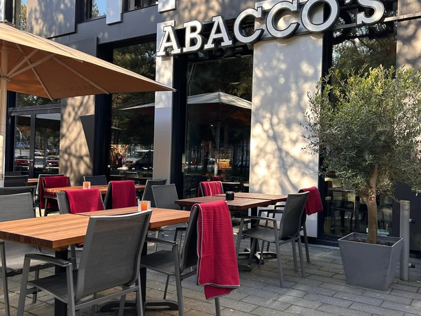 Abaccos Steakhouse Außen_3.jpg Außenbereich des Restaurants mit Tischen Stühlen, roten Decken und einem Sonnenschirm.