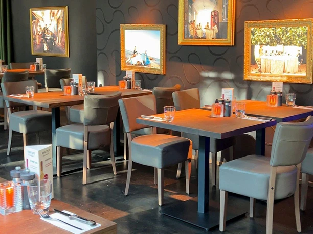 Innenraum des Restaurants mit Gemälden an den Wänden, gedeckten Tischen mit Stühlen in warmem Licht.