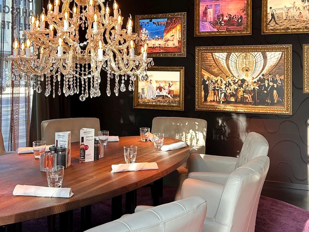 Edles Speisezimmer mit dunklen Wänden, an denen Bilder hängen, und einem großen Kronleuchter über einem ovalen Holztisch mit breiten Lederstühlen.