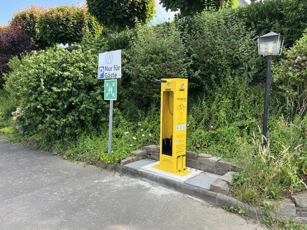 Radservice-Station Gelbe Fahrrad-Servicestation neben einem Parkplatzschild, umgeben von dichter grüner Vegetation.