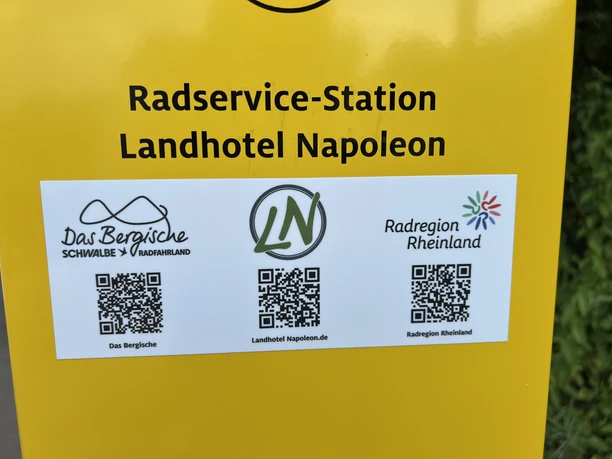 Radservice-Station Gelbes Schild mit Aufschrift "Radservice-Station Landhotel Napoleon" und drei QR-Codes darunter.