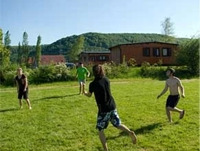 Campingplatz Axelsee - Freizeitgelände 4 Menschen, die auf dem Rasen miteinander Volleyball spielen, im Hintergrund Holzhütten und Bäume