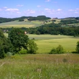 Landschaft mit grünen Feldern, sanften Hügeln und verstreuten Höfen unter blauem Himmel mit Wolken.