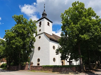 Weißes Kirchengebäude mit runden Fenstern und Turm, links- und rechtsseitig von Bäumen umgeben