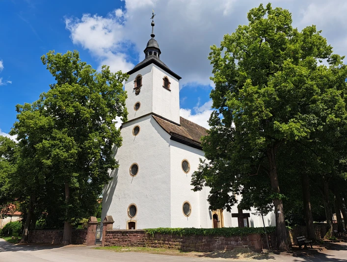 Kath. Pfarrkirche Hl. Familie und Hl. Stephanus in Wehrden Weißes Kirchengebäude mit runden Fenstern und Turm, links- und rechtsseitig von Bäumen umgeben