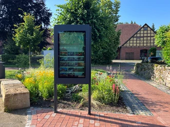 An outdoor information sign, surrounded by colorful flowers, with a historic half-timbered house in the background.