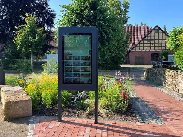 An outdoor information sign, surrounded by colorful flowers, with a historic half-timbered house in the background.