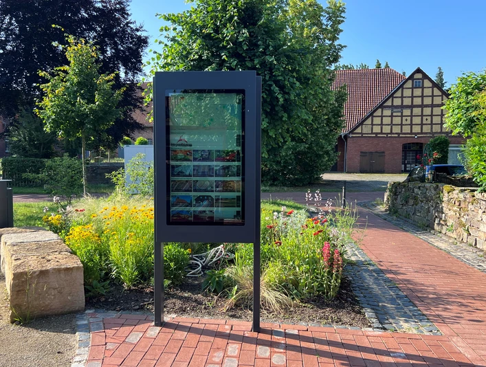 Infoterminal Winzlar Infotafel im Freien, von bunten Blumen umgeben, mit historischem Fachwerkhaus im Hintergrund.