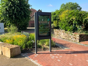 Infoterminal Winzlar Infoterminal Winzlar vor einer malerischen Kulisse mit Pflanzen und einem historischen Gebäude im Hintergrund.