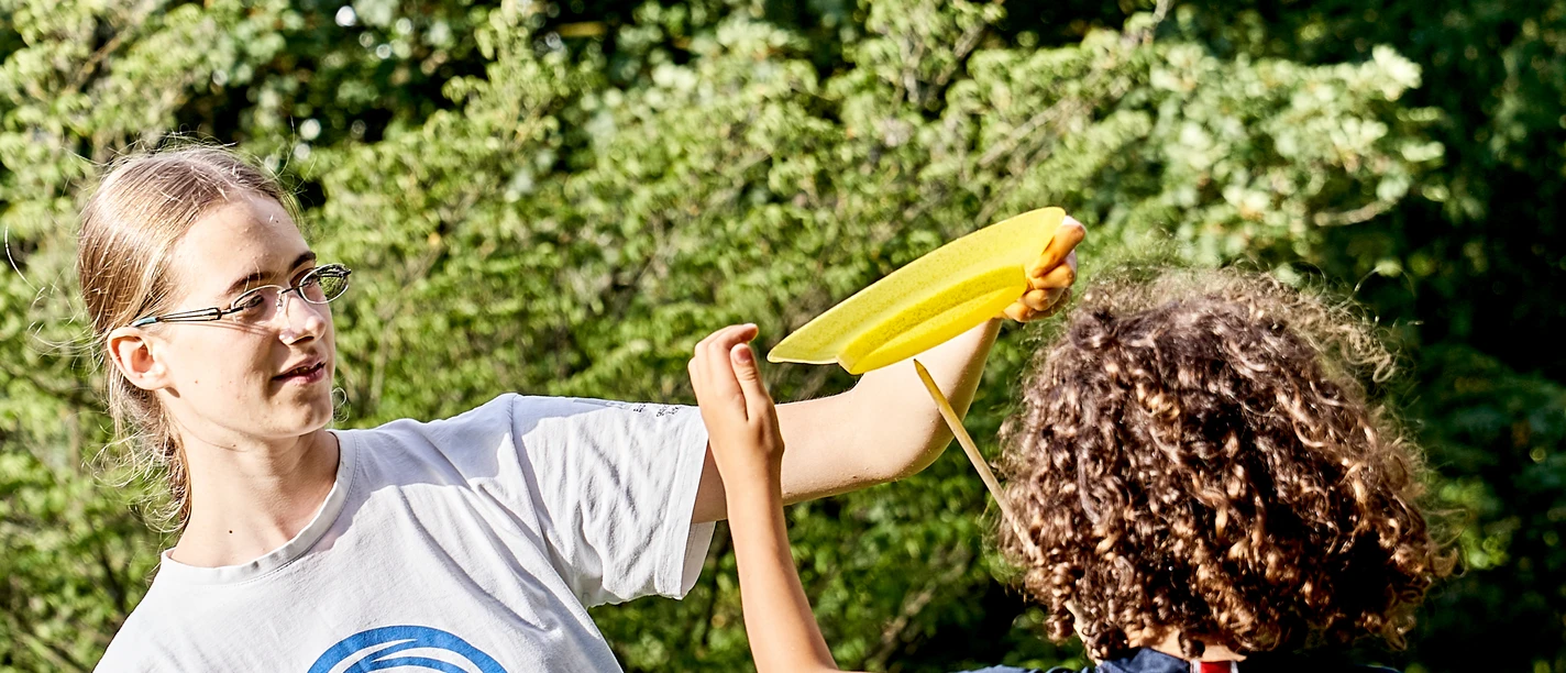 Hildener Sommer - Mitmachzirkus Erwachsene Person hilft einem Kind beim Jonglieren mit einem Teller im Park, umgeben von Bäumen.