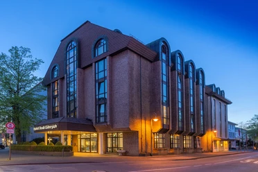Hotel Stadt Gütersloh Mehrstöckiges Stadthotel mit Backsteinfassade, großen Fensterbögen und beleuchtetem Eingang am Abend