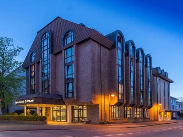 Hotel Stadt Gütersloh Mehrstöckiges Stadthotel mit Backsteinfassade, großen Fensterbögen und beleuchtetem Eingang am Abend
