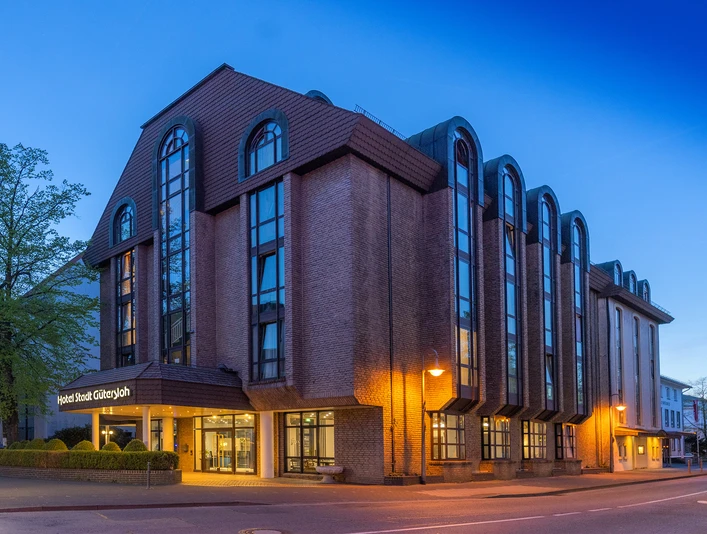 Hotel Stadt Gütersloh Mehrstöckiges Stadthotel mit Backsteinfassade, großen Fensterbögen und beleuchtetem Eingang am Abend