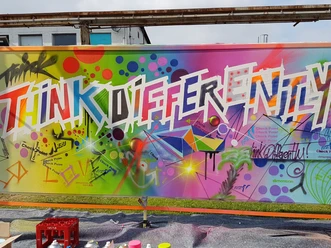 COLORI Events Graffiti wall with "Think Differently", in front of it colorful spray cans.
