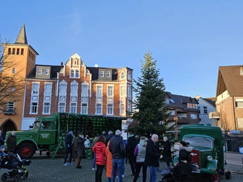 Ausschank unterm Weihnachtsbaum Menschen stehen in Reihen vor einem historischen Ausschankwagen der Marke Barre und einem Weihnachtsbaum auf einem gepflasterten Platz. Im Hintergrund sind Altbaugebäude bei klarem, blauem Himmel zu sehen.