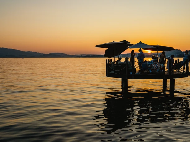 Guests on a lakeside terrace at sunset, with parasols and mountain silhouettes