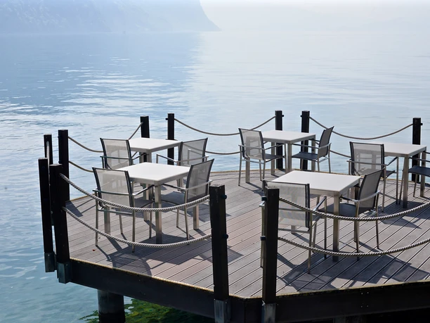 Lakeside restaurant terrace with tables and chairs overlooking calm water and hazy mountains