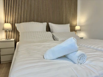 Schlafzimmer.JPEG Zwei frisch bezogene Betten mit weißen Laken, auf denen zwei gerollte Handtücher liegen.