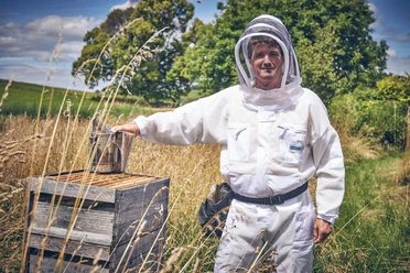 BienenFranz Ein Imker in Schutzkleidung steht neben einem Bienenstock auf einer Wiese.